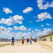 People walking along the coast next to colourful beach huts on a sunny day.