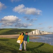 A couple walk together on a headland with white chalk cliffs in the background.