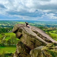 A seated woman waving from a hanging stone, overlooking a country panorama.