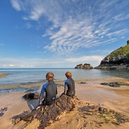 Back view of two young boys in wetsuits sitting on a rock looking out to sea with blue sky