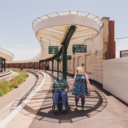 A woman walks with another woman who sits in a wheelchair at an old railway station