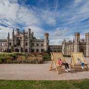 Man and woman holding ice creams in oversized deck chairs in manicured castle grounds and gardens.