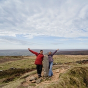 Un homme et une femme debout autour d'un grand rocher, les bras tendus, dans un paysage champêtre