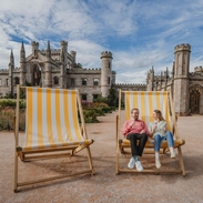 Man and woman holding ice creams in oversized deck chairs in manicured castle grounds and gardens.