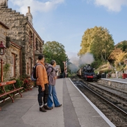 A man and woman standing on a train station platform as a train approaches in the background