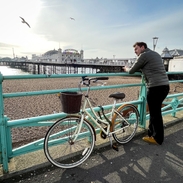 Junger Mann mit Fahrrad, der sich über das Geländer an der Strandpromenade von Brighton lehnt, mit Brighton Pier im Hintergrund