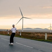 A male skateboarder enjoys the flat road surface to skate on in the Scottish Highlands, with wind turbines behind him