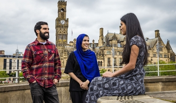 Two women and a man stand and talk in front of a town view